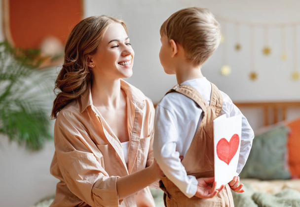 Boy hiding handmade greeting card with heart behind back while congratulating happy mother in bedroom - Foto, Bild