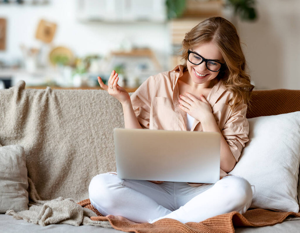 Jovem mulher positiva em roupas casuais e óculos sentados no sofá em casa e conversando em vídeo chat via netbook enquanto gesticulando e sorrindo - Foto, Imagem