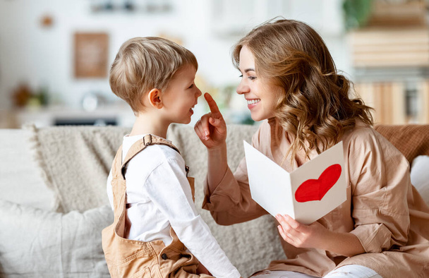 Cheerful mother hugging son and reading handmade greeting card with heart while resting on sofa during holiday celebration mothers day at home - Photo, Image