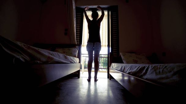 Жінка з силуету відкриває завісу в спальні і витягується після вставання вранці. - Фото, зображення