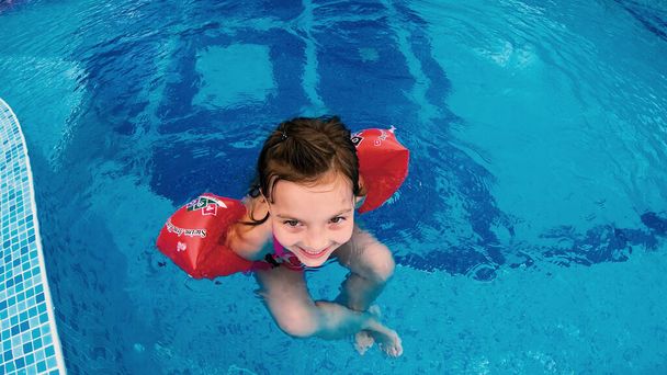 Happy little girl with inflatable arm bands relaxing in pool smiling - Фото, изображение