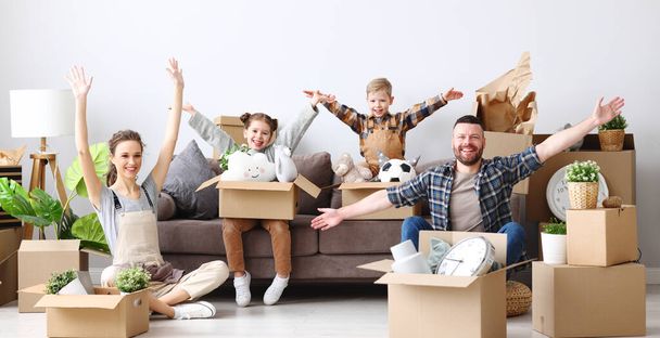 Zachwyceni rodzice i słodkie dzieci z podniesionymi ramionami gromadzące się na kanapie w salonie z rozpakowanymi pudełkami, patrząc przed kamerą i ciesząc się przeprowadzką w nowym mieszkaniu - Zdjęcie, obraz