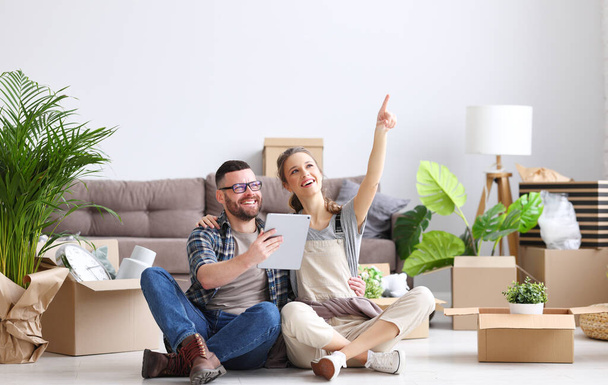 Fröhliches Paar sitzt mit Tablet und ausgepackten Kartons auf dem Boden, während es über die Dekoration der neuen Wohnung diskutiert und wegzeigt - Foto, Bild