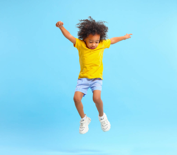 Full length of energetic little ethnic boy with Afro hairstyle in stylish casual outfit jumping with raised arms and looking down against blue background - Photo, Image