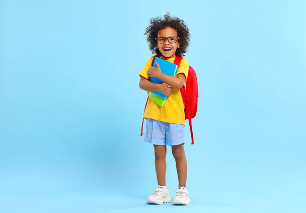 Full body of positive little African American boy with curly hair in casual clothes and eyeglasses smiling while standing against blue background with school backpack and copybooks - Foto, imagen