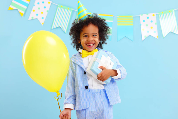 Cheerful little black boy with curly hair in stylish suit and party hat smiling while standing against blue background decorated with colorful flags with yellow balloon and present in hands - Photo, Image