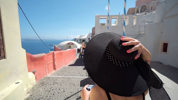 Elegant tourist woman with hat on travel vacation in Oia, Santorini, Greece. Happy young woman walking on narrow street iin Oia visiting the Greek island. - Photo, Image