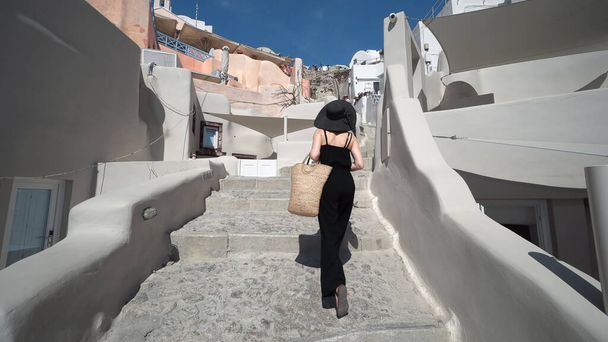 Elegant woman in black climbing stairs on Santorini. Young female is on summer vacation in Santorini. She is visiting famous tourist attraction in Oia - Foto, afbeelding