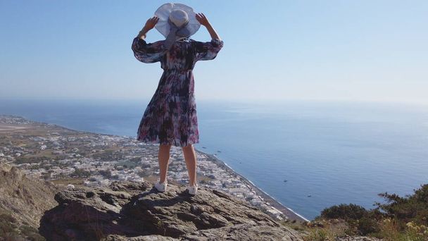 Happy woman tourist having fun swirling around in joy on Santorini. Panoramic view of Kamari from Ancient Thera mountain archeologicl site, slow motion - 写真・画像