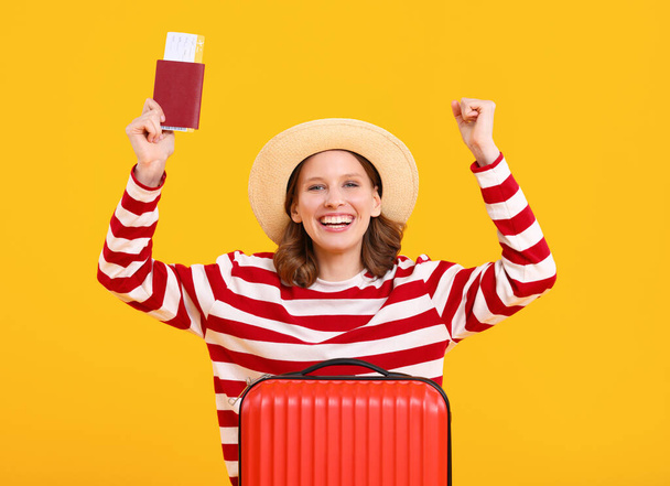 Jovem viajante alegre em camisa listrada e chapéu segurando passaporte com bilhete e mala e olhando para a câmera contra fundo amarelo brilhante - Foto, Imagem