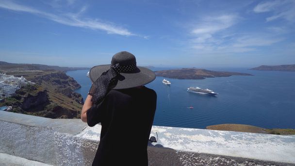A woman with hat using a public binocular that oversee the view of the Santorini Caldera including the volcanic island of Nea Kameni - Foto, afbeelding
