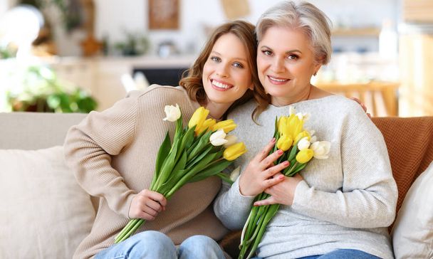 Positieve jonge dochter met senior moeder in casual kleding glimlachen en knuffelen terwijl zitten op de bank thuis met trossen van verse delicate tulpen tijdens de viering van de internationale Vrouwendag - Foto, afbeelding