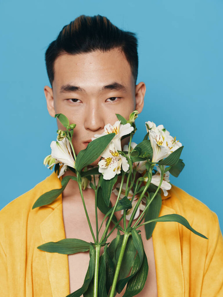 Cute Korean man in a yellow coat with a bouquet of white flowers on a blue background model - Foto, Bild
