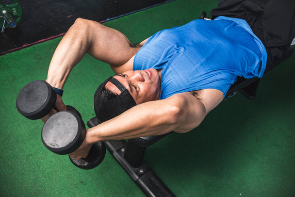 Un hombre asiático en forma hace un conjunto de extensiones de tríceps de pesa mentirosa. Entrenamiento Tríceps y brazos en el gimnasio. - Foto, Imagen