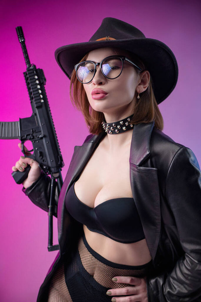 μια κομψή γυναίκα με μαύρα ρούχα και καπέλο ποζάρει σε φόντο νέον με αεροβόλο όπλο - Φωτογραφία, εικόνα
