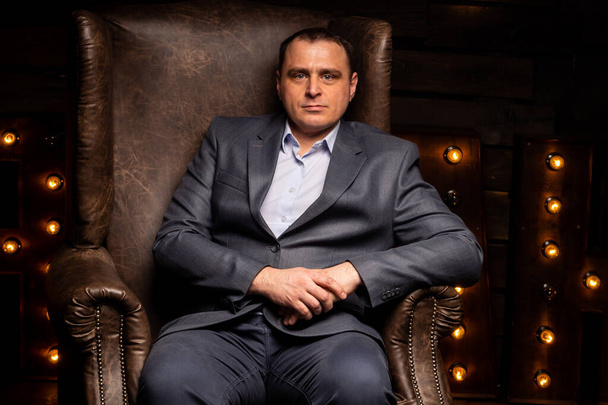 hombre de negocios guapo en un traje mira a la cámara en una silla de cuero. aislado sobre un fondo negro - Foto, Imagen
