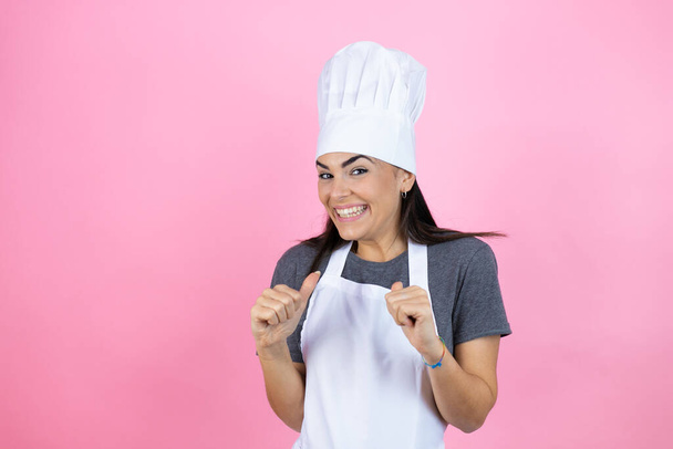 Молодая латиноамериканка в мундире пекаря на розовом фоне гордо и уверенно улыбается руками на бедре, счастливая и уверенная в успехе, придавая "успешный" вид. - Фото, изображение