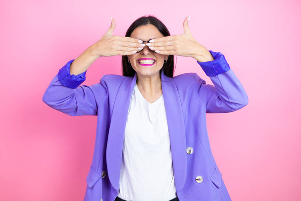 Joven mujer de negocios con chaqueta púrpura sobre fondo rosa cubriendo los ojos con las manos sonriendo alegre y divertido. Concepto ciego. - Foto, imagen