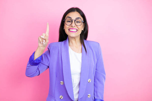 Mladá obchodní žena nosí fialovou bundu přes růžové pozadí ukazující a ukazující nahoru s prsty číslo jedna a zároveň se usmívá sebevědomě a šťastně - Fotografie, Obrázek
