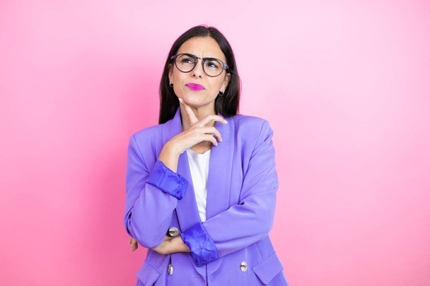 Young business woman wearing purple jacket over pink background serious face thinking about question with hand on chin, thoughtful about confusing idea - Photo, Image