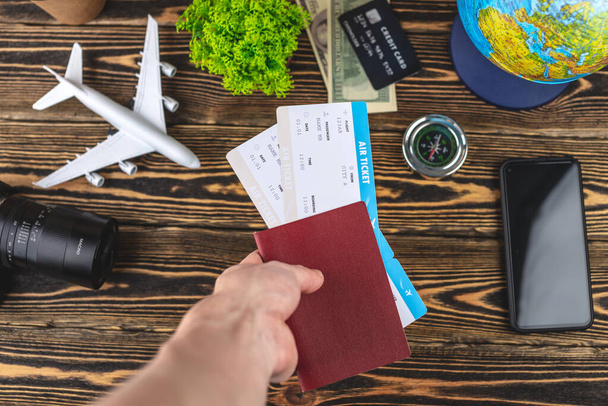 La mano sostiene un pasaporte y boletos de avión sobre una mesa de madera con suministros de viaje. Concepto de vacaciones y reservas. Vista superior. - Foto, imagen