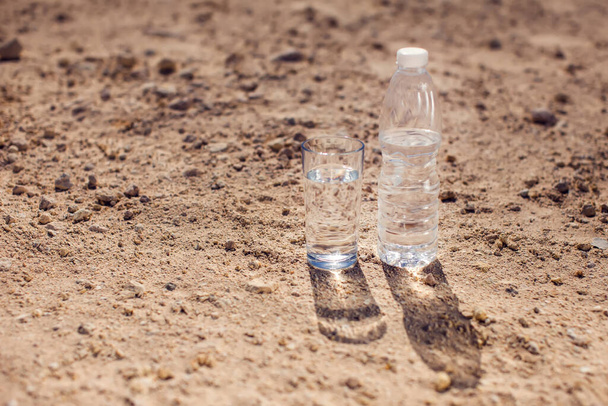 Çölde kum üzerinde cam ve bir şişe su. Susuzluk, sıcaklık ve kuraklık kavramı - Fotoğraf, Görsel