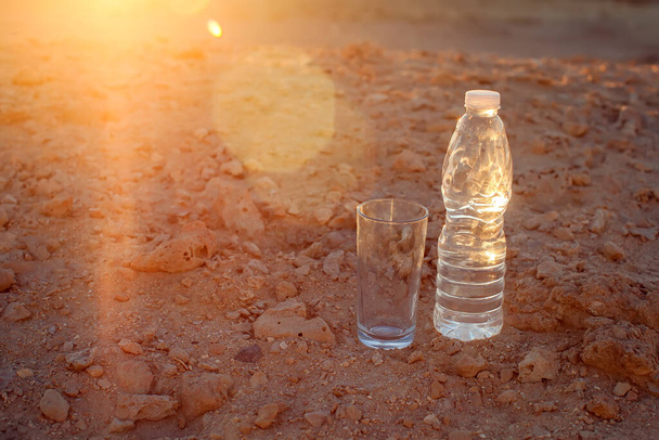 Verre et bouteille d'eau sur sable dans le désert. Soif, chaleur et sécheresse concept - Photo, image