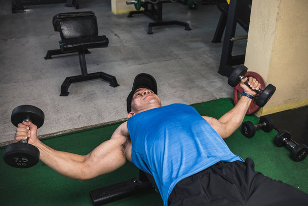 Un hombre asiático con una camiseta azul hace un juego de flys en el pecho de una mancuerna en un banco plano. Pectorales de entrenamiento y parte superior del cuerpo en el gimnasio. - Foto, imagen