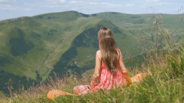 Jovem mulher em vestido vermelho sentado no campo gramado em um dia ventoso nas montanhas de verão desfrutando de vista da natureza. - Filmagem, Vídeo