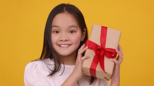 Linda chica asiática sacudiendo caja de regalo, tratando de coger lo que está dentro, fondo naranja, cámara lenta - Imágenes, Vídeo