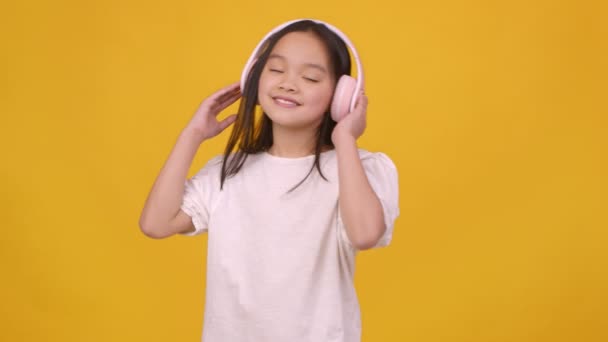 Schattig klein aziatisch meisje genieten van klassieke muziek en dansen, het dragen van draadloze headset, oranje achtergrond, slow motion - Video