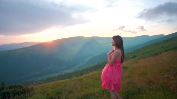 Fiatal nő piros ruhában áll a füves réten egy szeles őszi hegyekben, felemeli a kezét, élvezi a természet látványát.. - Felvétel, videó