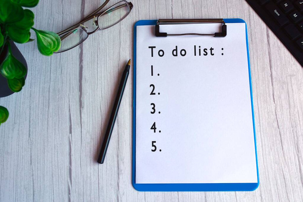 Για να κάνετε λίστα κειμένου σε μπλε πίνακα κλιπ με γυαλιά, στυλό, φυτό και πληκτρολόγιο σε ξύλινο γραφείο.  - Φωτογραφία, εικόνα