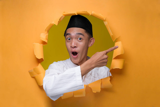 Aziatische moslim tiener man poseert door gescheurd geel papieren gat met geschokt gezicht, het dragen van moslim doek met schedel cap, wijzend een kopieerruimte om iets te presenteren. Reclame voor of verkoop van producten. - Foto, afbeelding