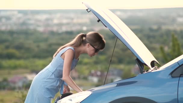 Stranded giovane donna autista in piedi vicino a una macchina rotta con spuntato cofano ispezionare il suo motore del veicolo. - Filmati, video