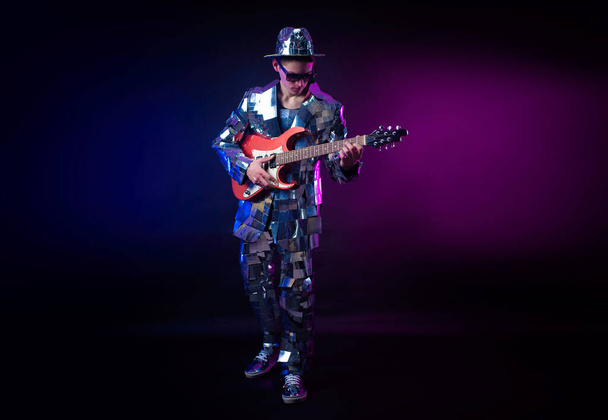a male artist in a mirrored suit and hat with an electric guitar on a dark background - Photo, Image