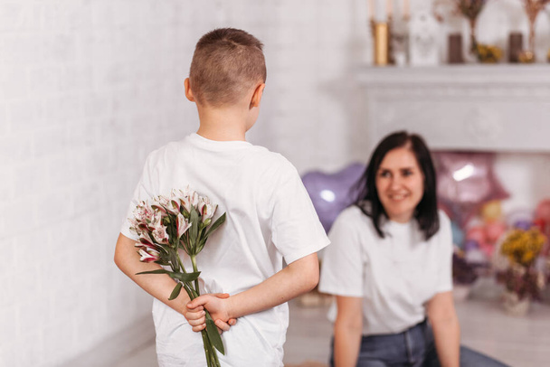 Le garçon tient des fleurs derrière son dos et félicite sa mère - Photo, image