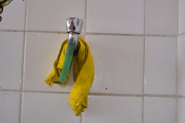 Metal faucet and piece of green hose on it. Yellow cleaning towel hanging on it with white faience background. - Photo, Image
