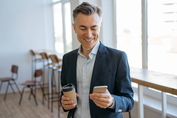 Kendine güvenen olgun bir adam elinde akıllı telefon ve bir fincan kahve tutuyor, haberleri okuyor, internetten iletişim kuruyor. Gülümseyen işadamı modern ofiste mobil uygulama kullanıyor.  - Fotoğraf, Görsel
