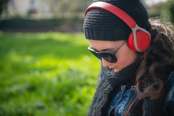 Jovem sentada em um banco vestindo fone de ouvido vermelho ouvir música. - Foto, Imagem