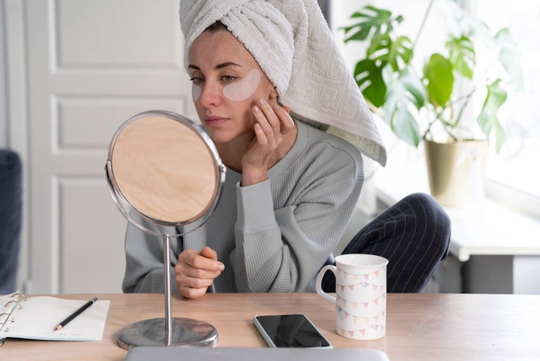 Vrouw met handdoek op het hoofd het aanbrengen van vlekken onder de ogen verrijkt met collageen, vitamine E, vermindert de tekenen van veroudering, helpt roodachtige oogwallen, kijken in de spiegel. Gezicht huidverzorging schoonheid thuis.  - Foto, afbeelding