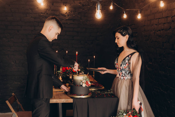 Свадьба красивой пары в черном платье, букет цвета марсалы, за праздничным столом со свечами и тортом у черной стены - Фото, изображение