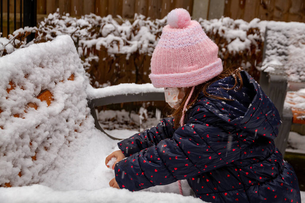 Ritratto di una graziosa bambina in cappello rosa che gioca nella neve indossando una maschera facciale - Foto, immagini