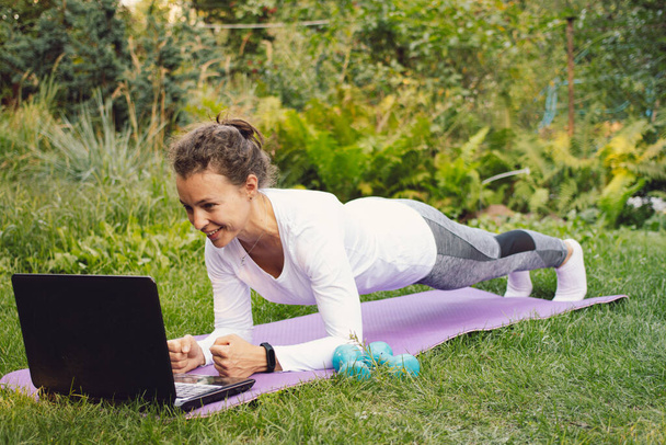 Bilgisayarında fitness egzersizleri olan ve yeşil parkta spor minderiyle yoga tahtası yapan mutlu formda bir kadın. İnternet bağlantısı üzerinden antrenör talimatlarını takiben evde spor yapmak - Fotoğraf, Görsel