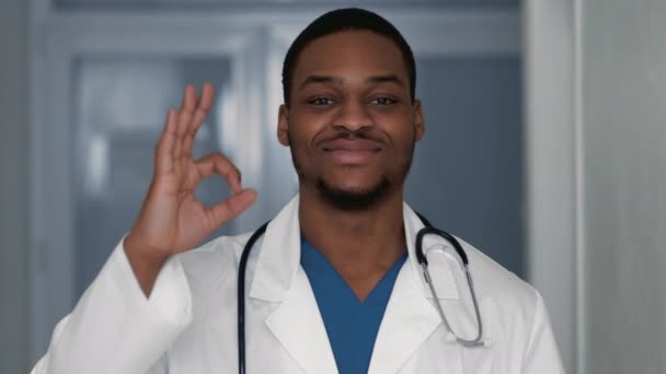 Positieve Afro-Amerikaanse mannelijke arts toont goed gebaar en brede glimlach op camera, kliniek interieur, slow motion - Video