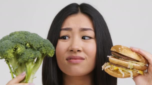 aziatische dame kiezen ongezonde hamburger in plaats van broccoli, witte achtergrond - Video