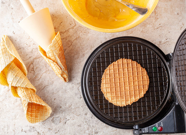 Flat lay image of non stick waffle cone maker to make homemade ice cream cones. Fresh made hand rolled cones are lying on marble countertop. Ingredients are in the bowl. - Photo, Image