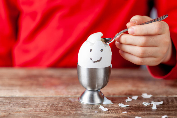 Een klein kind eet zacht gekookt humpty dumpty ei geplaatst in ei beker op een houten ontbijttafel. Ze gebruikt lepel om het ei uit zijn schelp te eten. Ze heeft een lachend gezicht op de schelp getekend..  - Foto, afbeelding