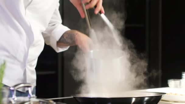 Joven chef profesional en la cocina prepara pasta asiática. hombre utiliza pinzas de olla caliente para poner fideos en wok con verduras fritas para una deliciosa comida gourmet preparada en una cocina casera. Cocina asiática - Metraje, vídeo