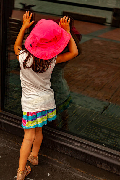 Una niña linda con ropa de verano se está presionando a sí misma a la puerta de cristal de un edificio tratando de ver lo que está pasando en el interior. Una imagen aislada de un niño preescolar curioso apoyado en la puerta - Foto, imagen
